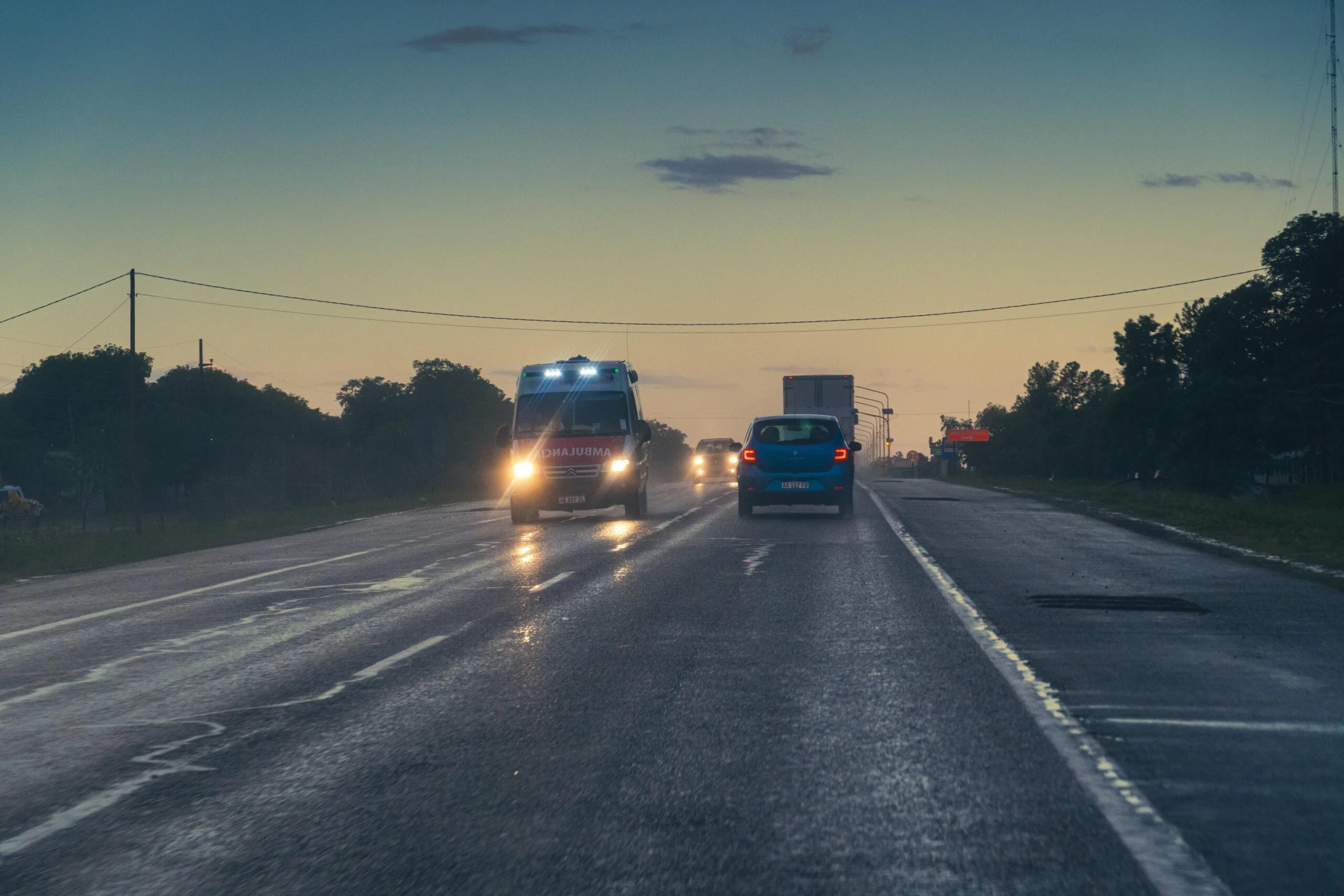Ambulance with its headlights and blue lights on, driving along a highway at dusk